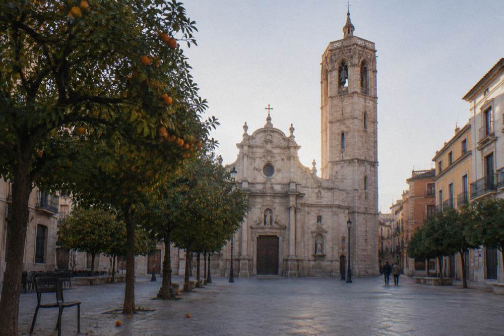 An old town square in Valencia with a church facade and orange trees along the edge