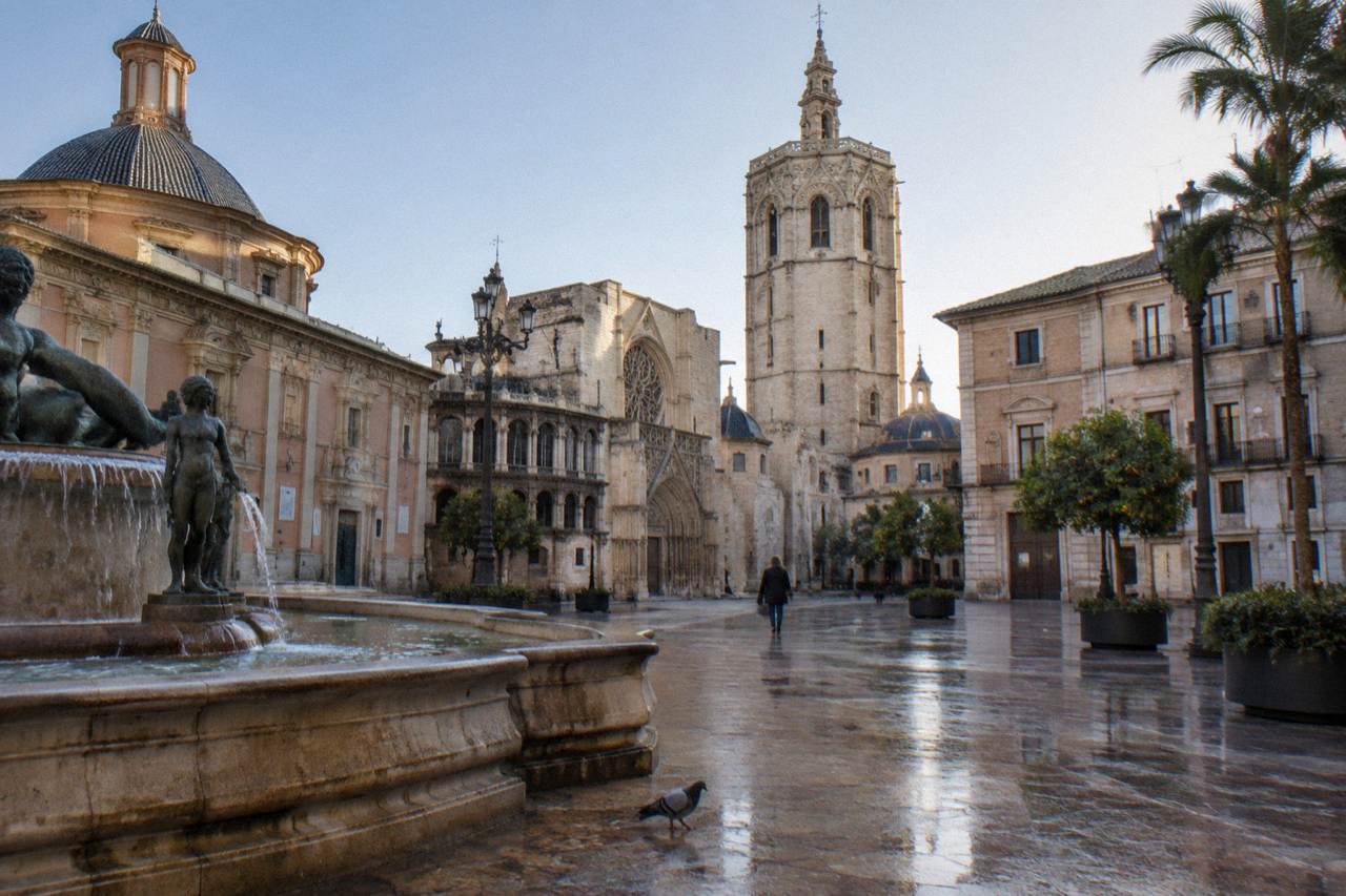 A sunlit plaza in Valencia with ornate historic buildings and a stone tower in the background.