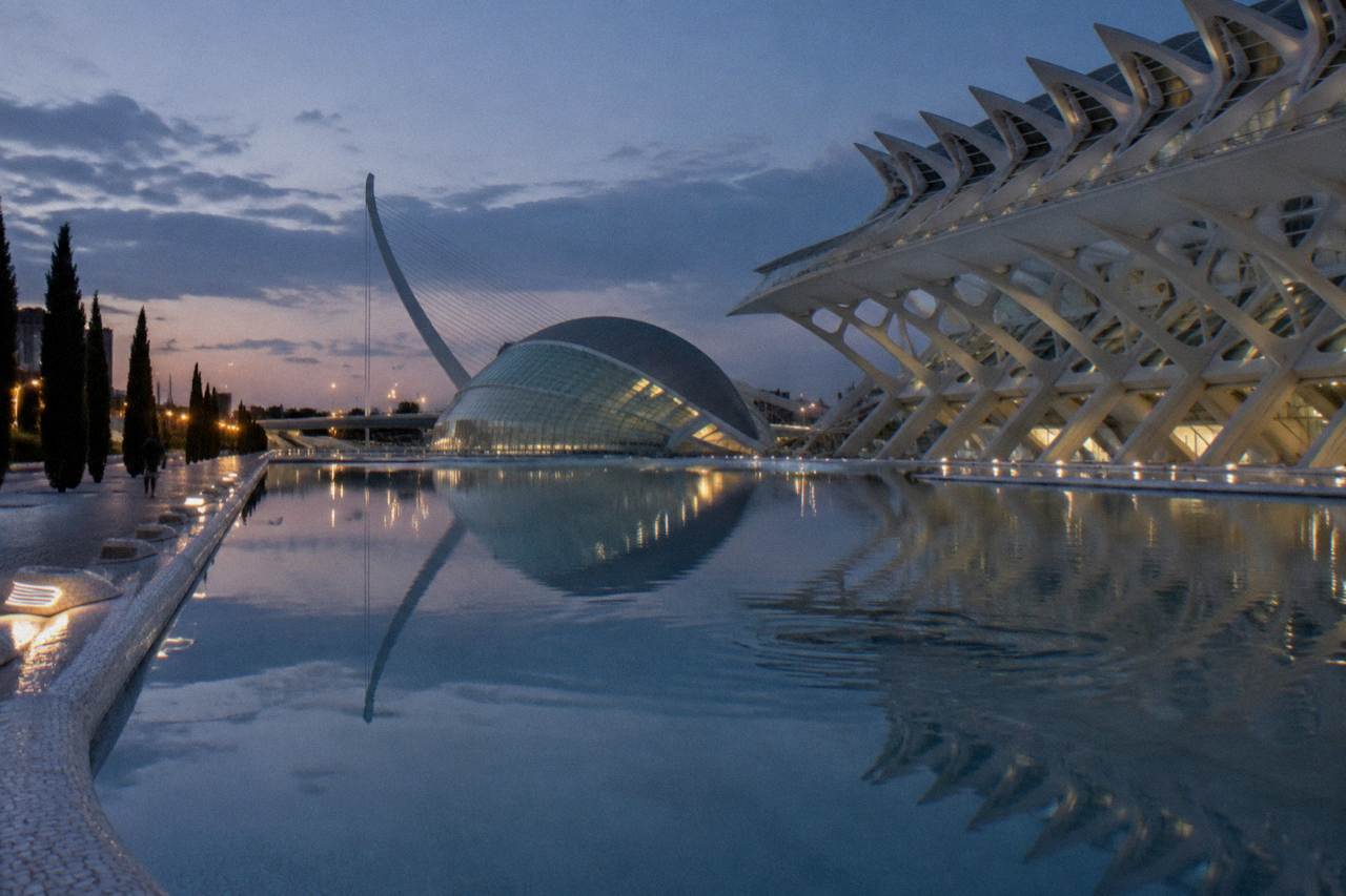 White futuristic buildings of the City of Arts and Sciences reflected in a wide pool under a clear sky