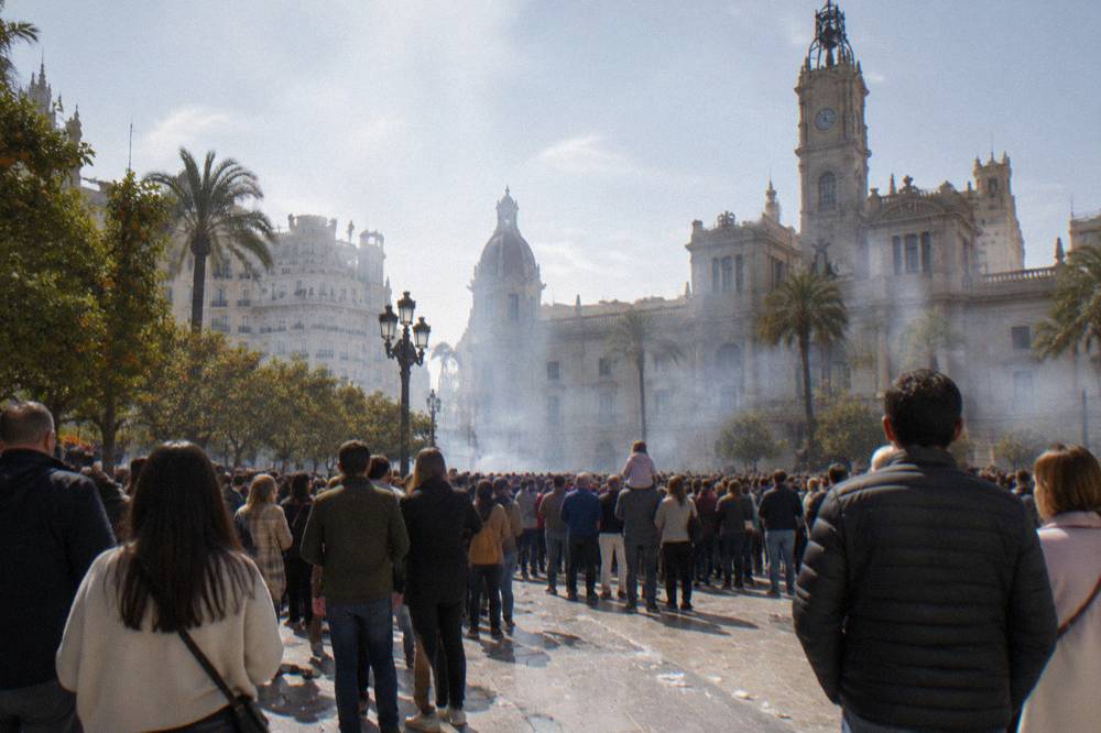 A wide city square filled with a dense crowd under a midday sky with smoke drifting above the rooftops