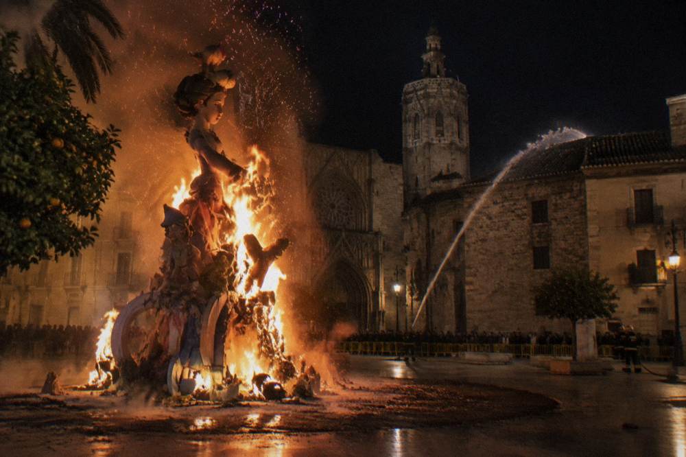 A large fallas monument ablaze at night with firefighters spraying water on nearby rooftops