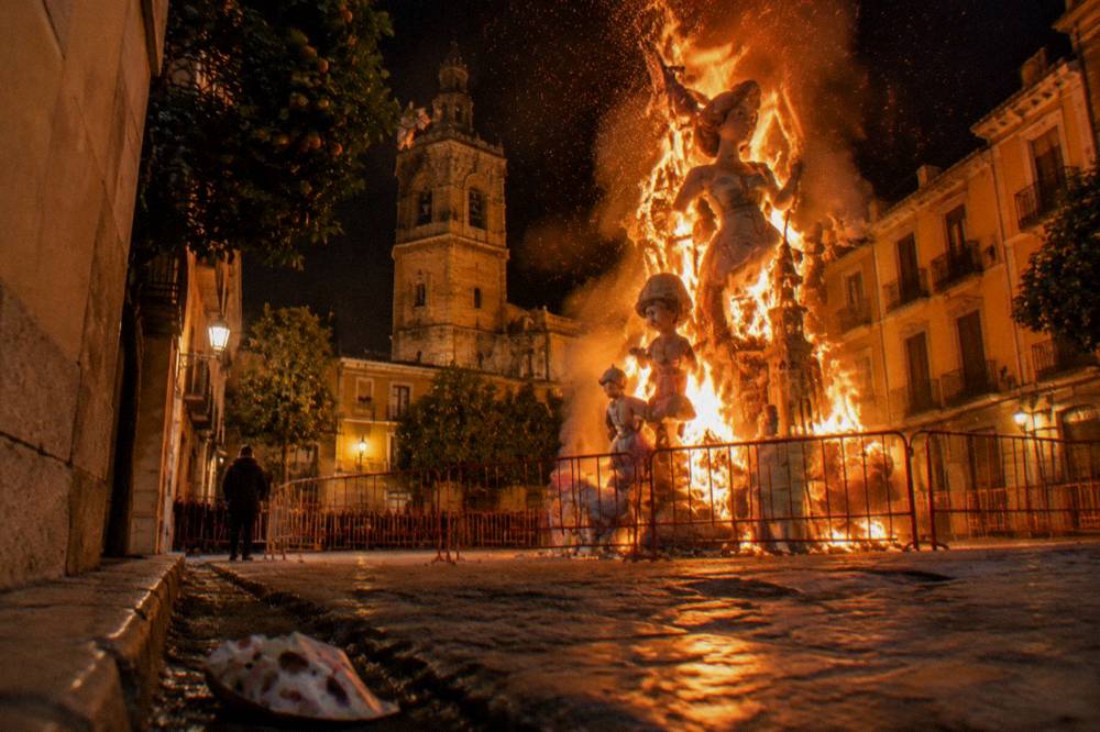 A falla monument engulfed in flames at night with sparks rising above a Valencia street