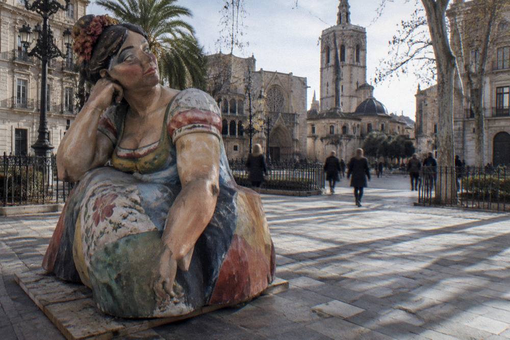 A painted falla figure at street level with a crowd walking past it in a Valencia square