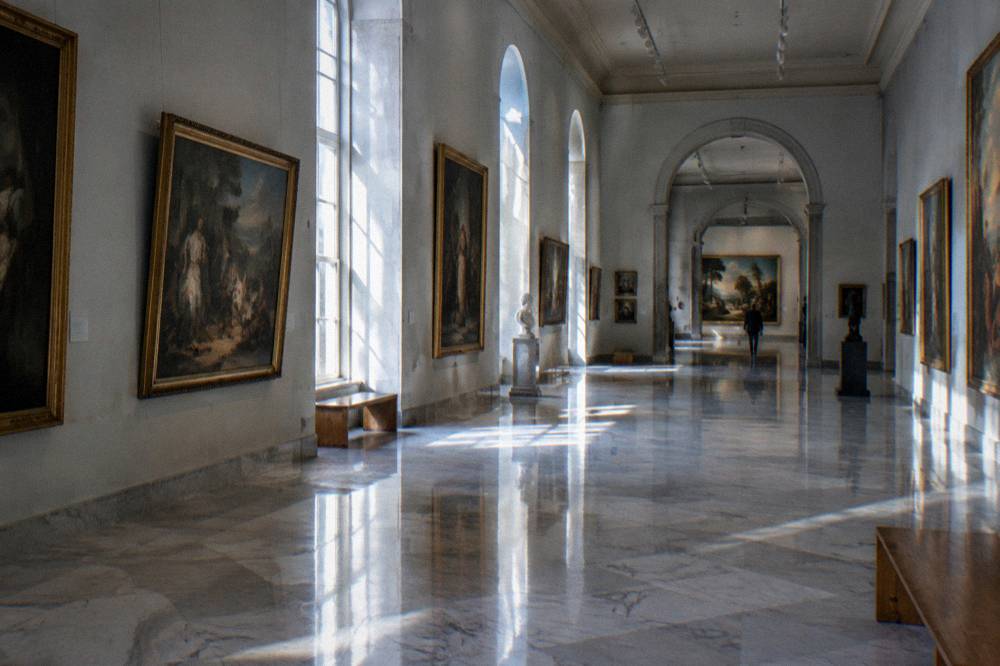 Sunlit gallery hall with marble floors and framed paintings along both walls