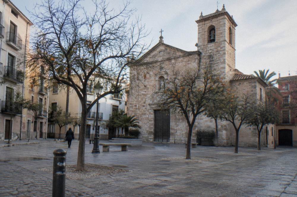 The stone facade and bell tower of a neighbourhood church above a small square with trees