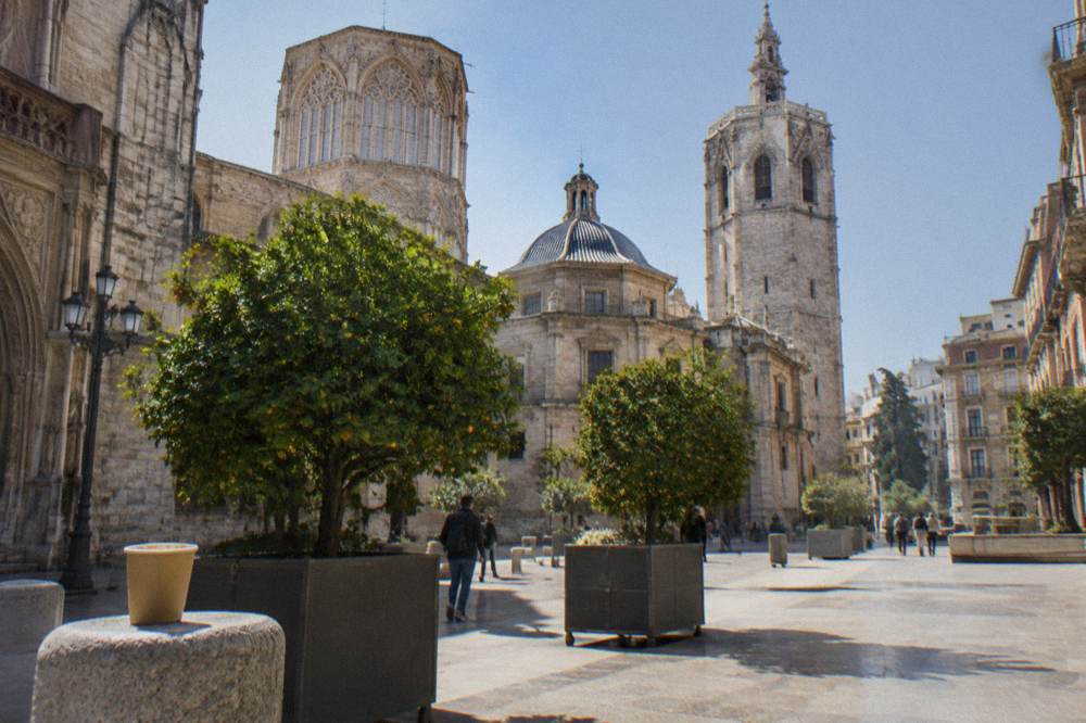Stone bell tower rising beside a cathedral with a domed roof in a bright plaza