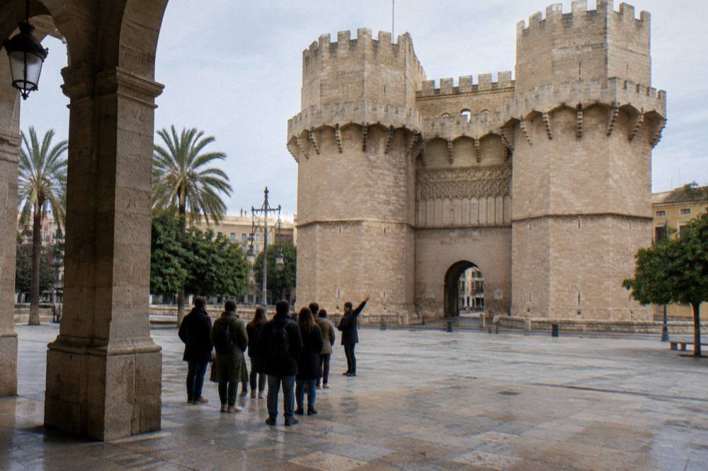 A walking guide gesturing toward a stone gate tower while a small group stands on the plaza